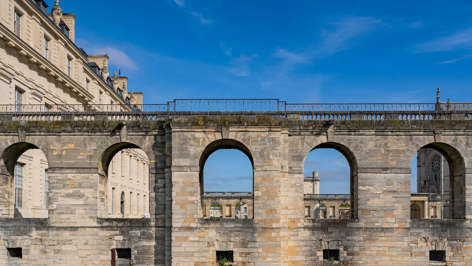 Arcaded stone walls inside the Château de Vincennes under a blue sky, with the Sainte-Chapelle's rose window framed in the far arch